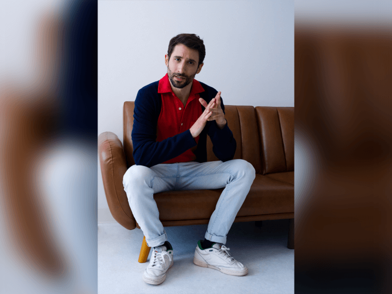 Actor Andy Dispensa sitting on a brown leather couch indoors with his arms resting on his knees, hands crossing.