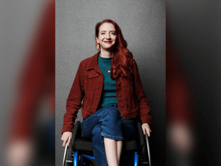 Actor Carey Cox proudly sitting in her wheelchair indoors, wearing a burgundy cardigan over a green shirt and jeans.