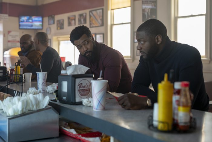 Isaiah Mustafa as John Sampson, Aldis Hodge as Alex Cross siting t a diner counter in burgundy and black turtlenecks.