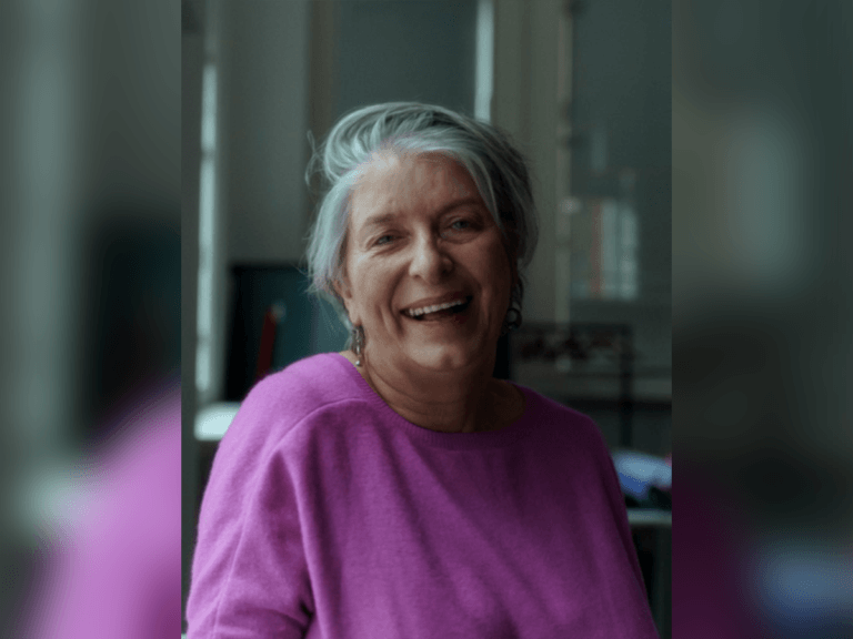 Maureen Hughes laughing and smiling indoors wearing a purple sweater.