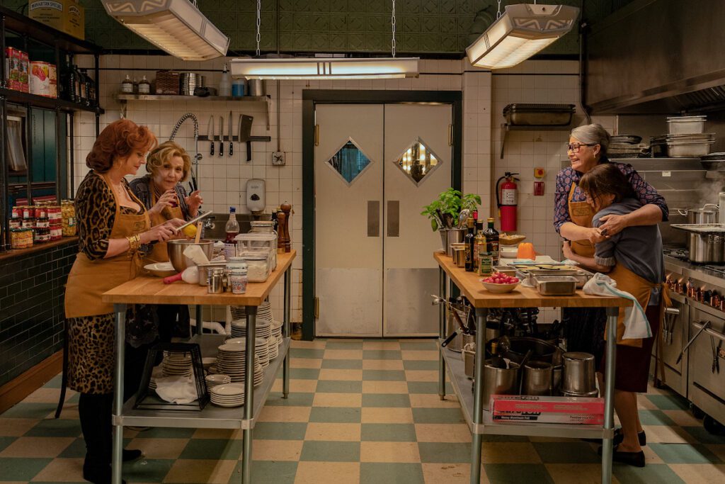 Susan Sarandon, Brenda Vaccaro, Lorraine Bracco and Talia Shire cooking on opposite ends of the kitchen of an Italian restaurant wearing brown aprons.