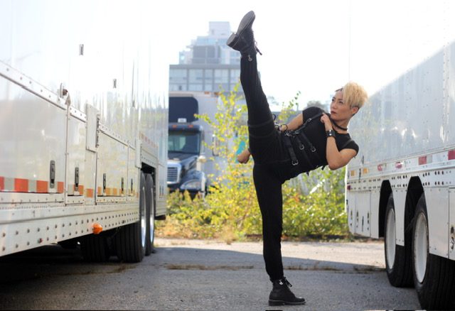 Juju Chan Szeto practicing her martial arts, doing a split kick, dressed in black mercenary gear between movie trailers.
