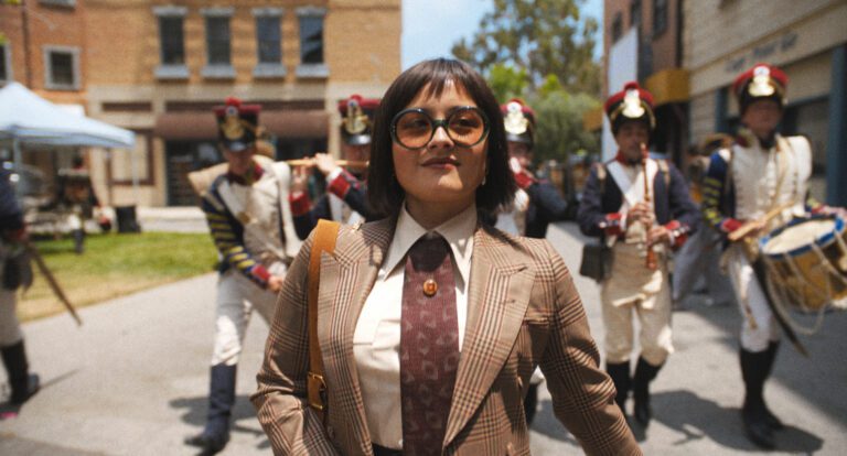 Chase Sui Wonders smiling confidently and walking through a film studio lot with a marching band behind her.