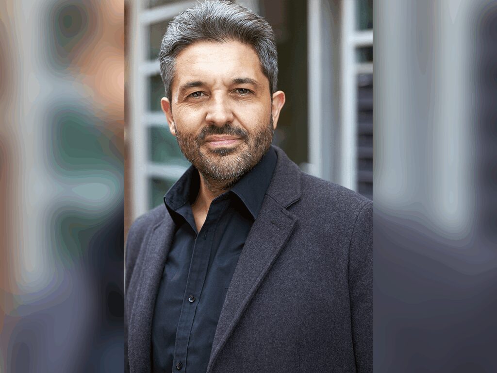 Val Athanassiou looking sharp in a black dress shirt and a charcoal blazer outside.
