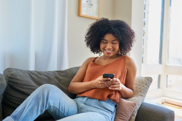 Young lack woman in jeans and a burnt orange top on her phone while casually laying on the couch.