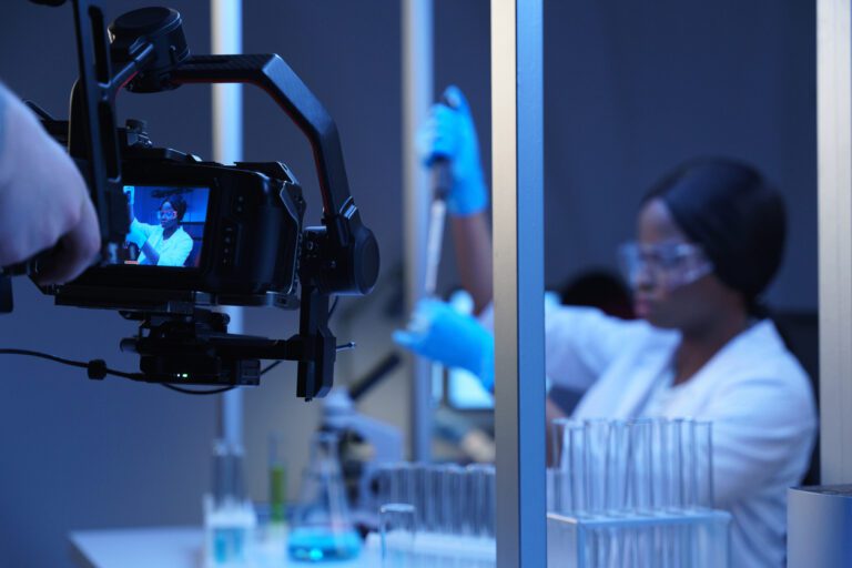Actress portraying a doctor filming a scene in a lab, surrounded by lab tech and a nearby camera.