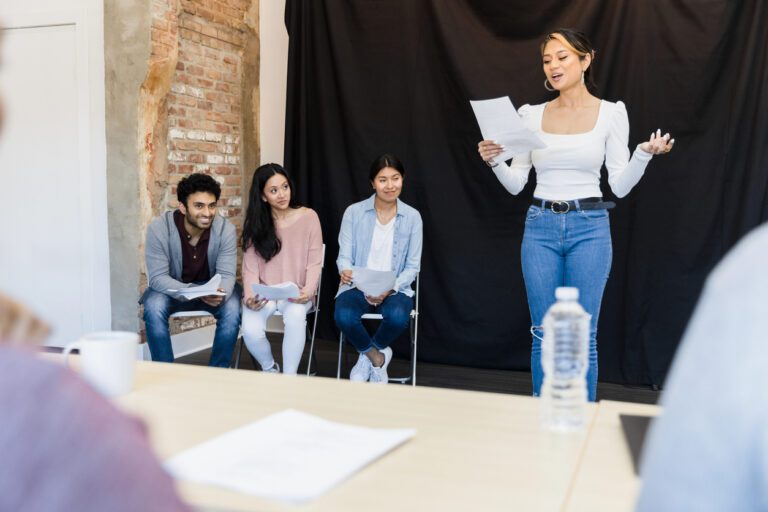 Asian actress, script in-hand, auditioning for casting directors in a brick room with a black curtain and diverse group of her fellow actors.