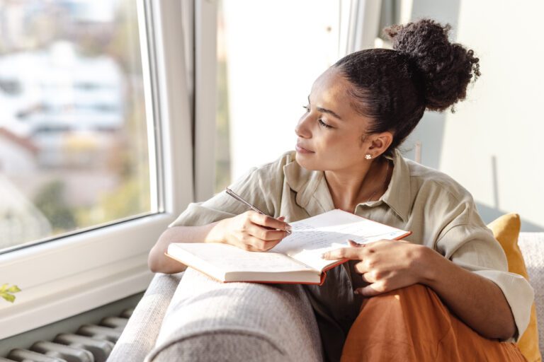Young biracial woman looking out the window on a sunny day while journaling on the couch.