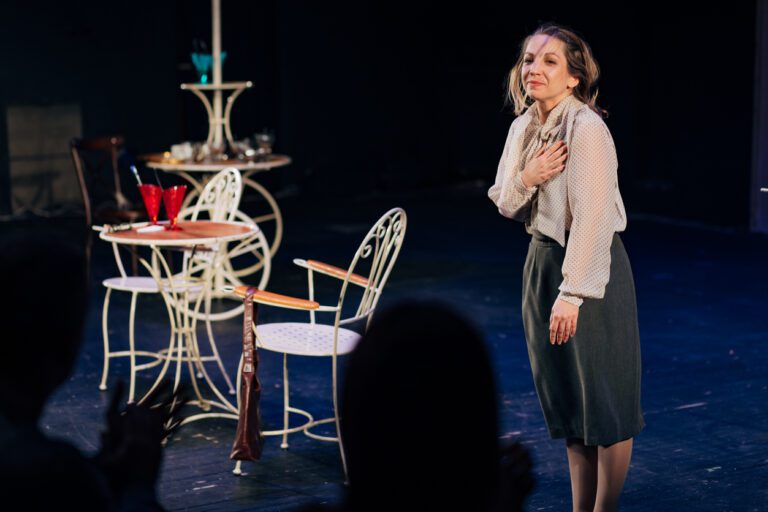 An actress on stage, set with a coffee table, chairs and red glasses, gives a heartfelt goodbye/bow to her audience as the performance ends.