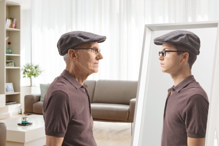 Old man wearing a cap and brown shirt looking into a mirror seeing his younger self wearing the same clothes.