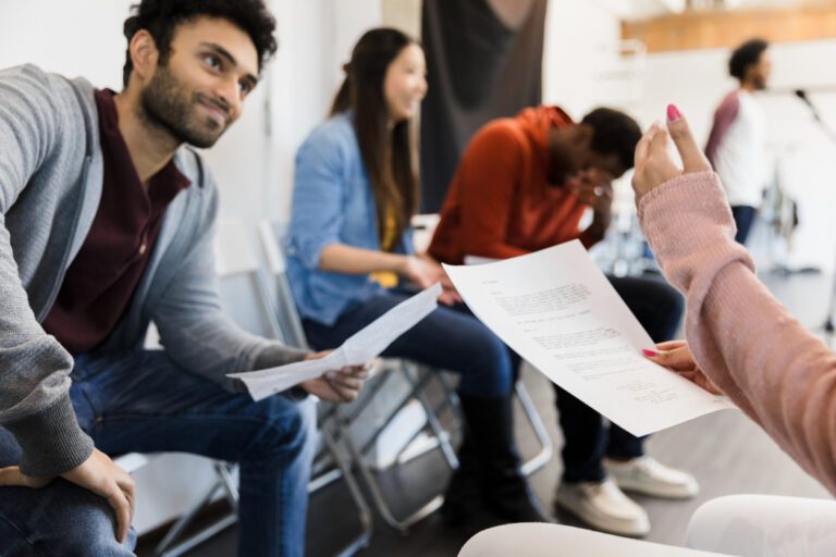 Diverse group of actors at an in-person audition, waiting their turns as one auditions for casting directors. Some look stressed, holding sides.