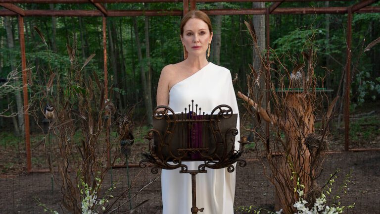 Julianne Moore dressed in Greek siren robes standing in an unkempt garden reading music on a rusty stand.