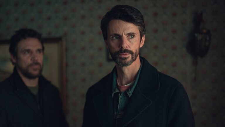 Matthew Goode inside a dimly lit apartment investigating, wearing a dark jacket.