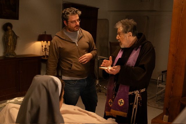 Al Pacino in priest robes, an actress playing a nun and Director David Midell in a church bedroom area for a movie scene.