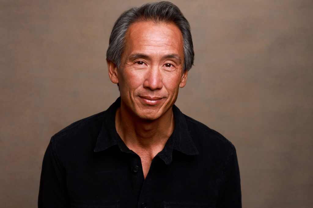 Actor Tinpo Lee standing up and smiling in a black sweater.