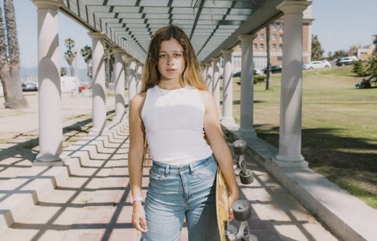 Pauline Sala walking through a park outside under a structure, skateboard in hand.