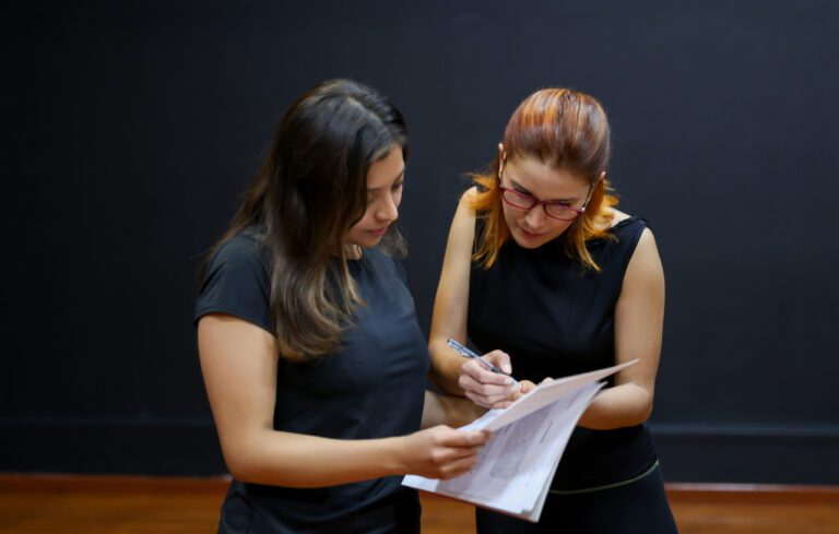 Two female actors in drama class wearing black shirts and black jeans going over a script.