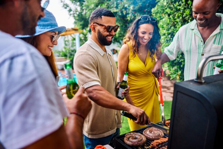 Diverse friend group hanging out in summertime grilling sausages and drinking beers at a park, watching the grill master.