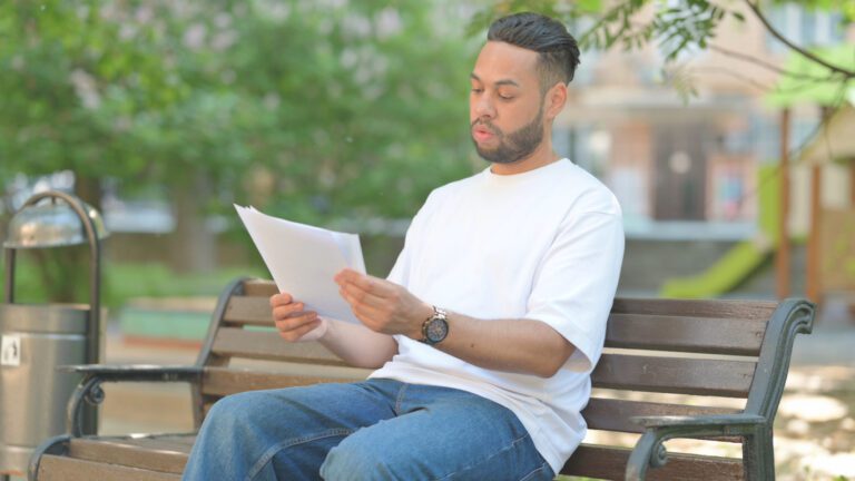 Young Spanish man in jeans and a white t-shirt reading a contract outside on a park bench.
