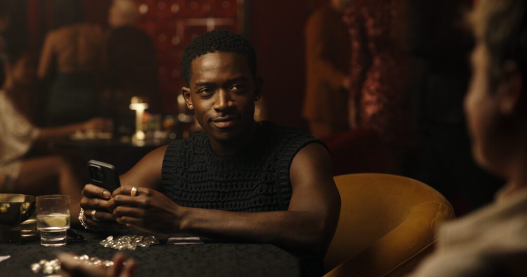 Damson Idris on his phone, talking to Brad Pitt, in the booth of a fancy restaurant in a cutoff black shirt.