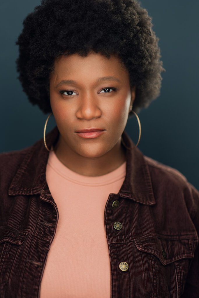 Actor Celine Milien smiling in front of a dark gray background wearing a burgundy denim jacket and a pink shirt.
