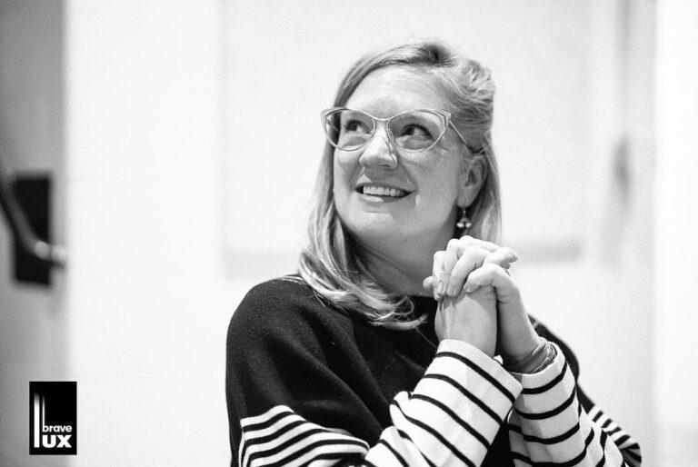Casting director Becca McCracken in a black sweater with striped sleeves holding her hands in prayer and smiling inside. A black and white filter is applied.