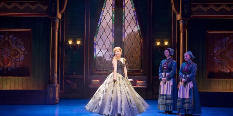 Laura Dawkes as ‘Anna’ smiling on a Norwegian-inspired hall stage set in ‘Frozen the Musical’
