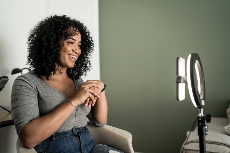 Spanish actor with curly hair sitting at home in a grey shirt and jeans filming on her phone being held by a ring light.