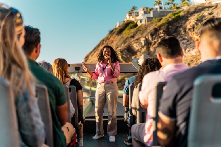 Female actor moonlighting as a summer tour guide on a bus by a cliffside house on a sunny day.