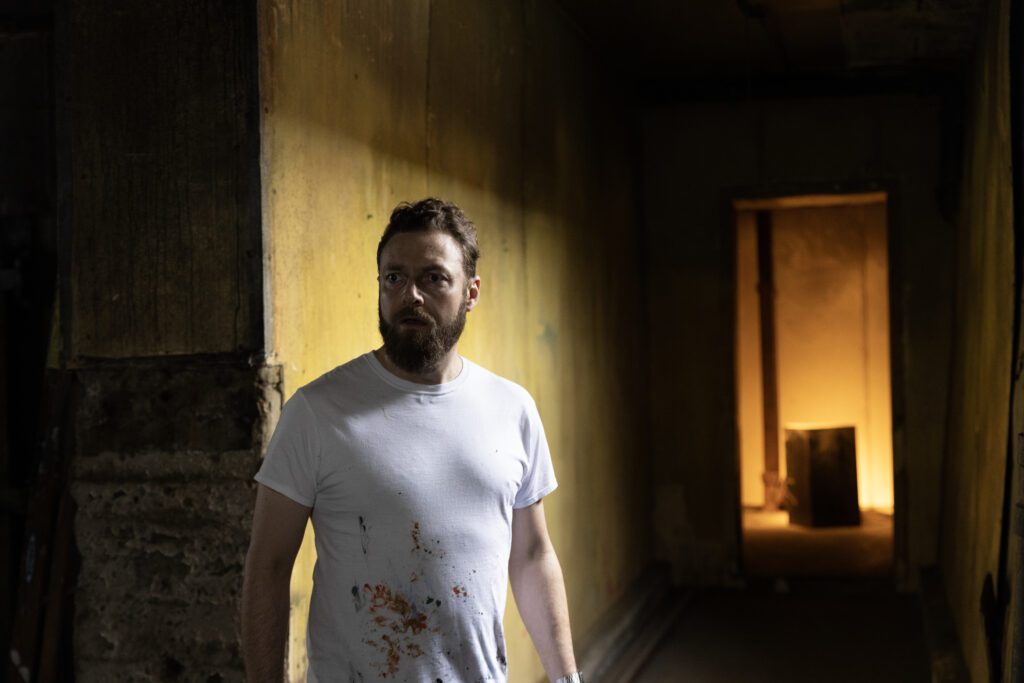 Ross Marquand walking down a creepy hallway, his white t-shirt is bloodstained.