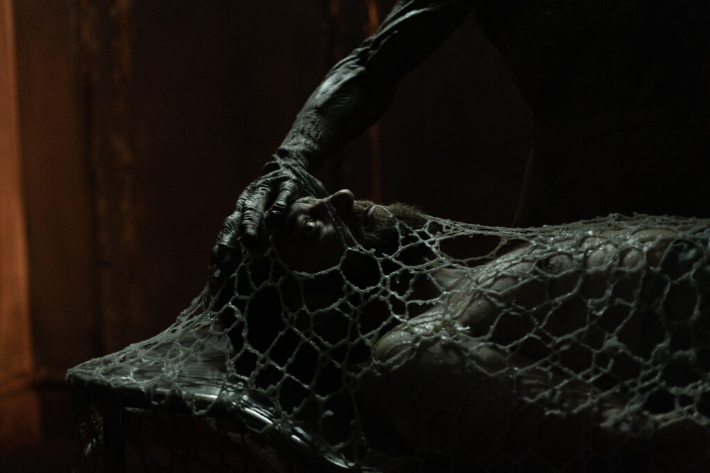 Ross Marquand lying flat in a diark area covered in a coccoon with a n alien hand over his head.