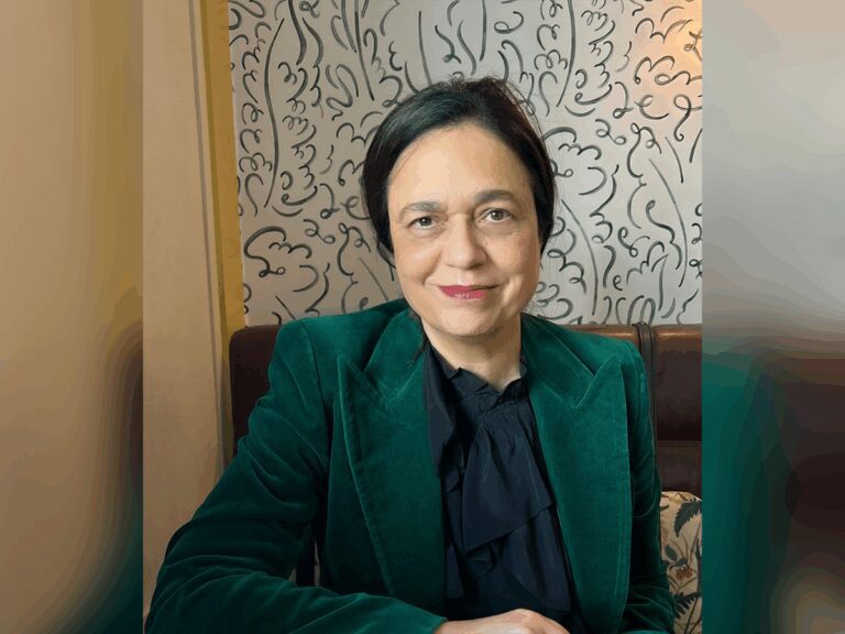 Casting director Jina Jay sitting in a chair by a doodle painting wearing a green blazer and black shirt.