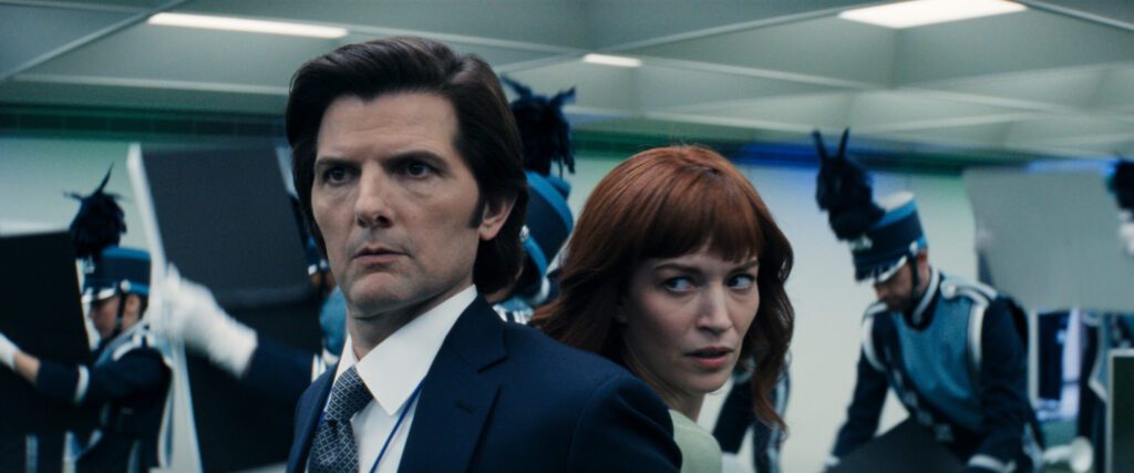 Severance stars Adam Scott and Britt Lower back-to back wearing professional work attire looking tense in an office room full of marching band soldiers.