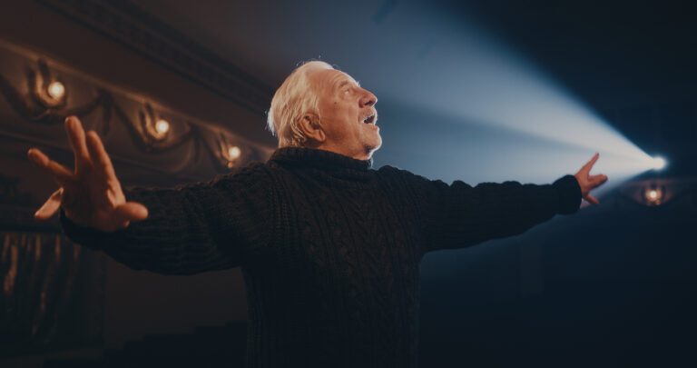 Older actor in sweater on stage during a theater performance making a grand gesture.