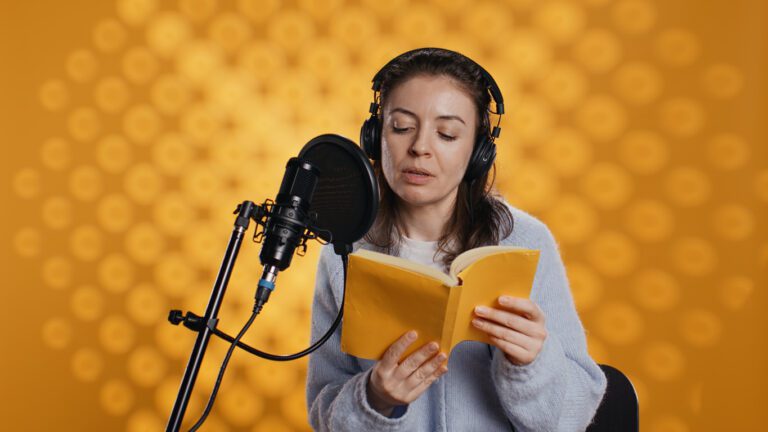 Audiobook narrator wearing headphones, reading aloud from book into microphone.