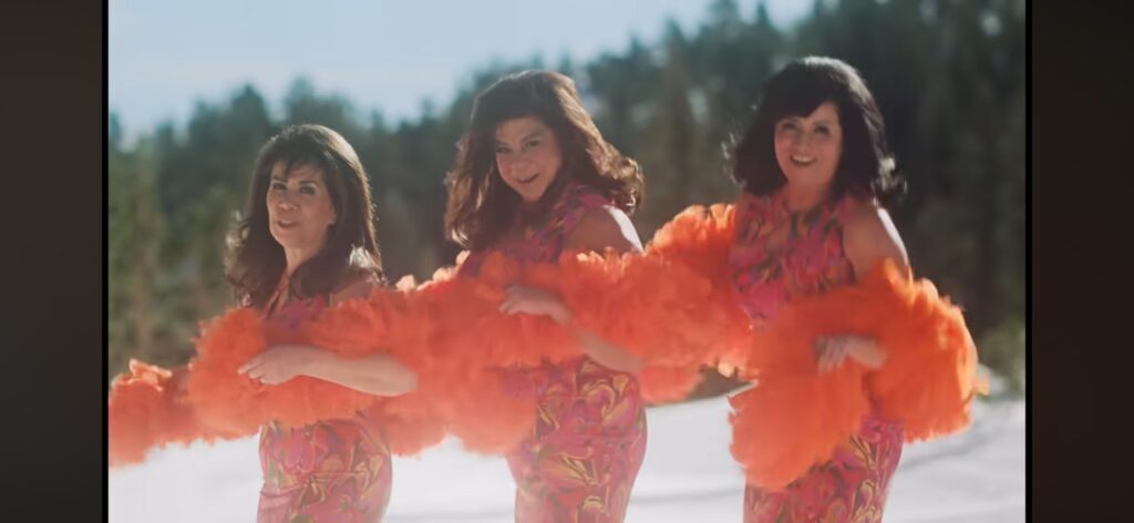 Stephanie Jones (center) and two other women dressed as older Spanish women wearing orange boas and pink outfits outside the cold mountains.