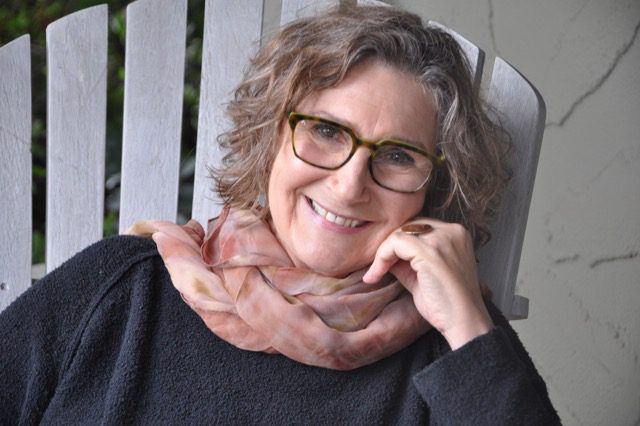 Retired casting director Jane Jenkins sitting on a white chair outside in a black sweater and pink scarf, hand on her chin.