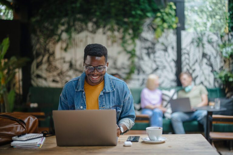 Young black man with glasses wearing a denim jacket and yellow shirt working on his laptop n a cafe.